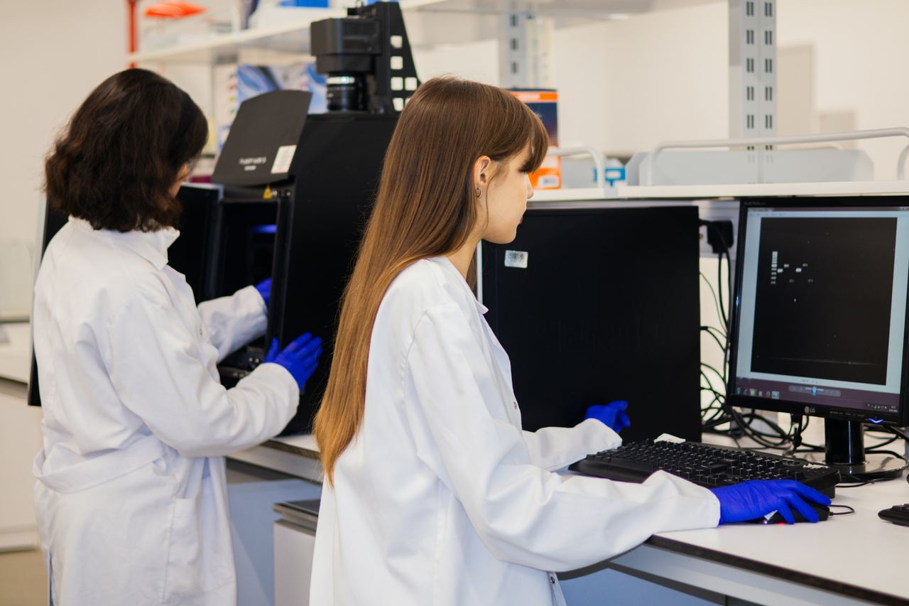Duas pesquisadoras de jaleco e luvas azuis num laboratório, uma delas digitando no teclado diante do monitor
